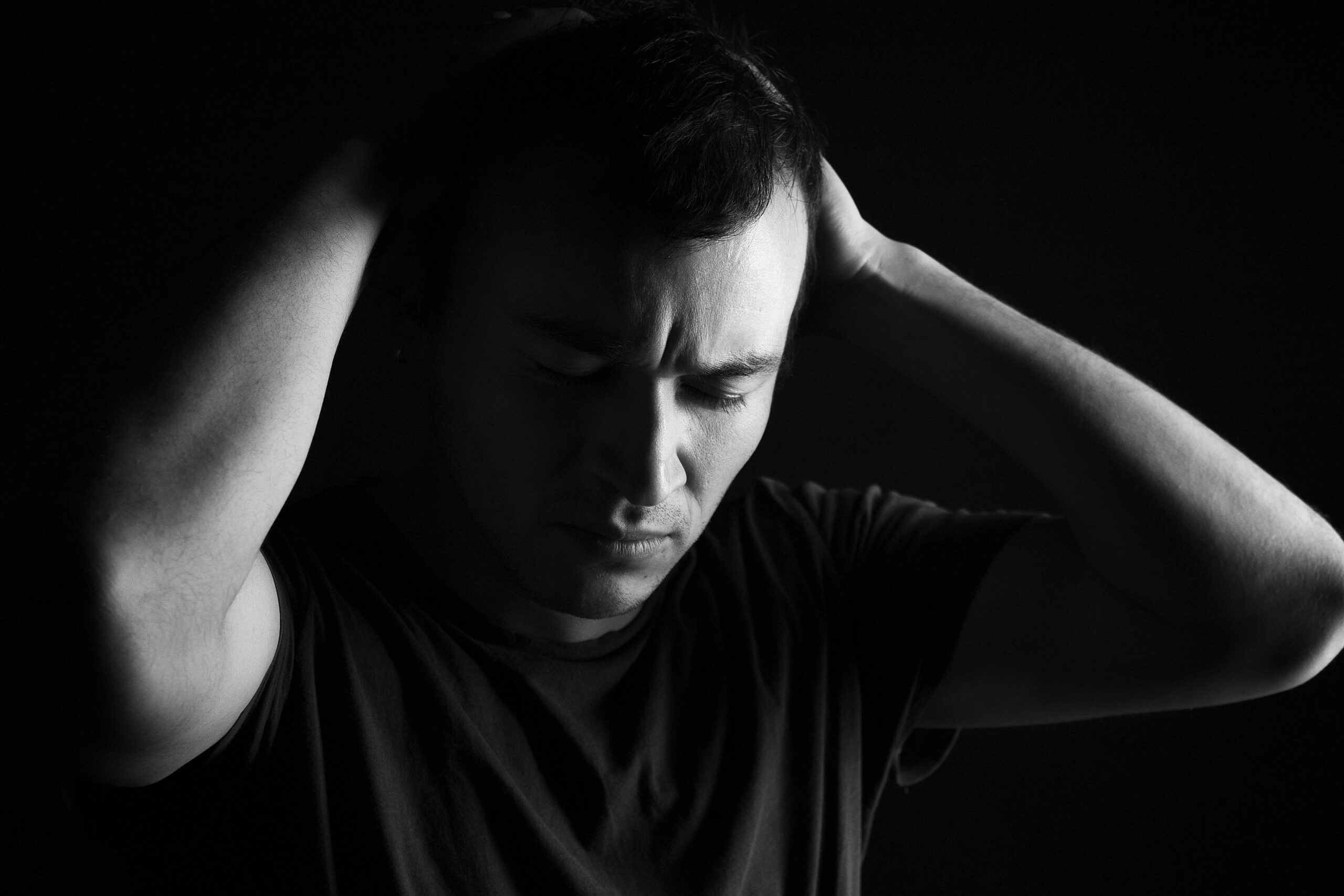 Man in grayscale with eyes closed, gripping his head with both hands, showing distress against a dark background.